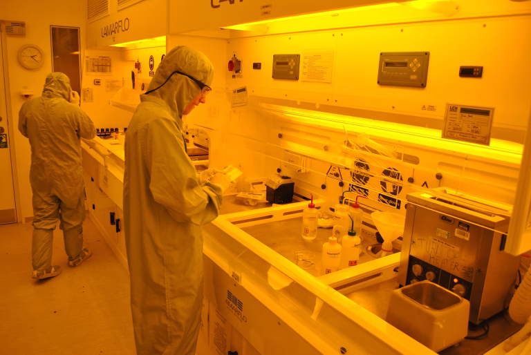 Technicians in cleanroom suits working at photolithography stations under yellow safelight
